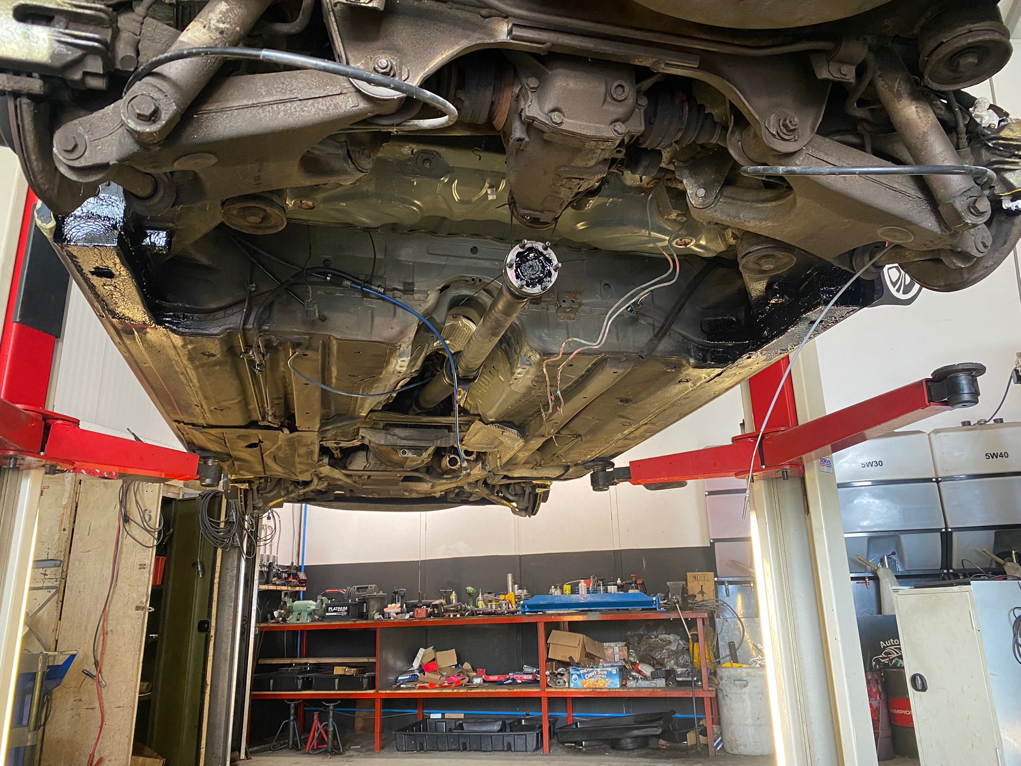 Onderkant van een auto op een tweepostsbrug, met werkplaatsrekken op de achtergrond